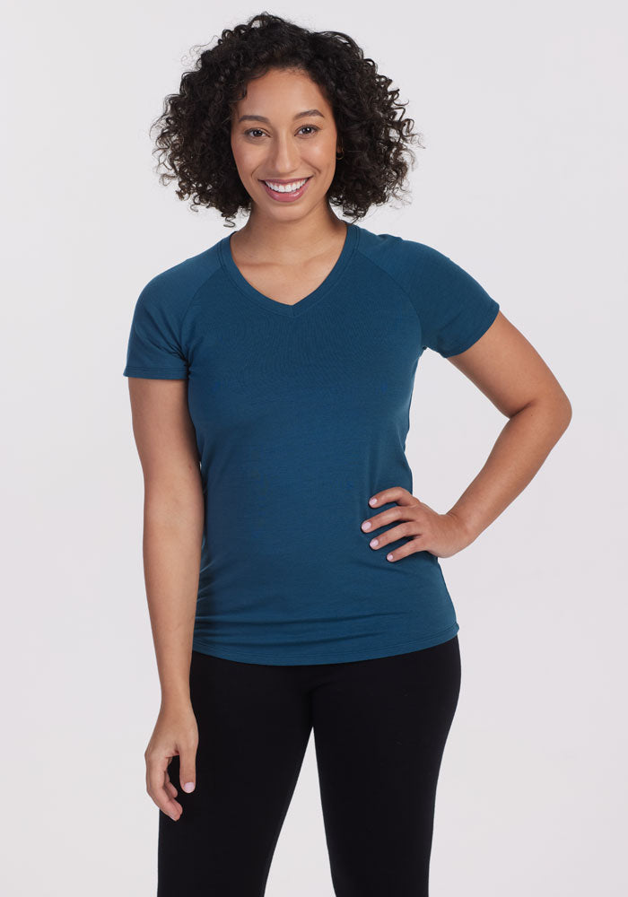 A person with curly hair is smiling, wearing the 'Mia V Neck' in Real Teal by Woolx and black pants. They stand against a plain white background, confident in their odor-free style. 