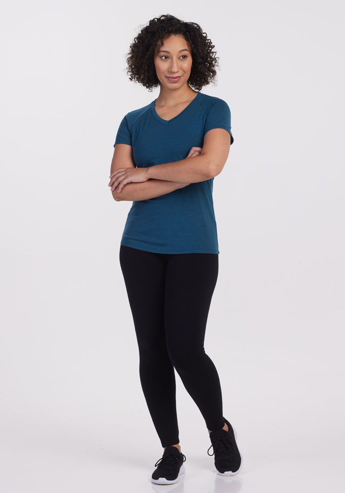 A person with curly hair stands confidently with crossed arms, wearing the Woolx Mia V Neck in Real Teal along with black leggings and black sneakers, set against a plain white background.