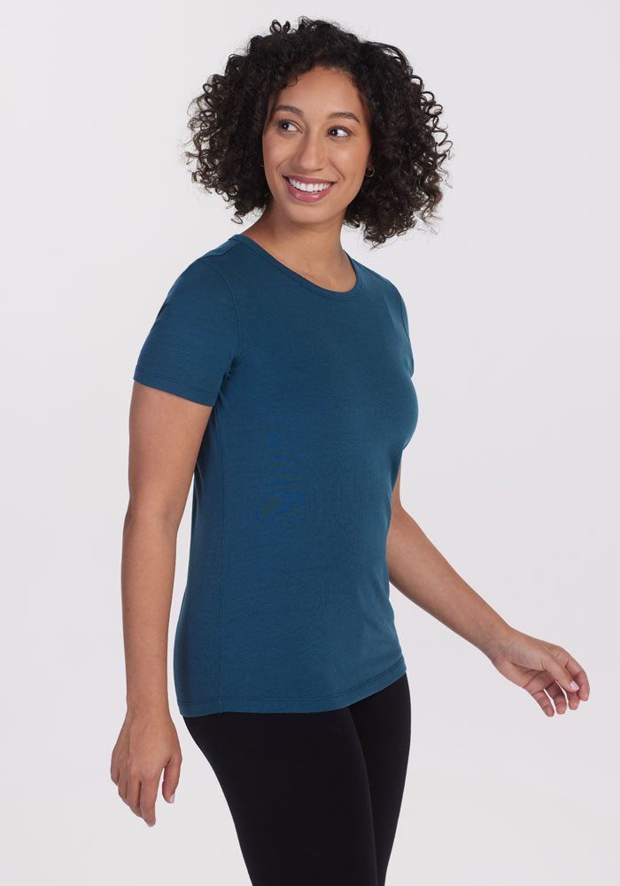 A person with curly hair, dressed in the Woolx Liza Crew Neck Tee in Real Teal and black pants, is smiling and looking to the side against a plain white background. 