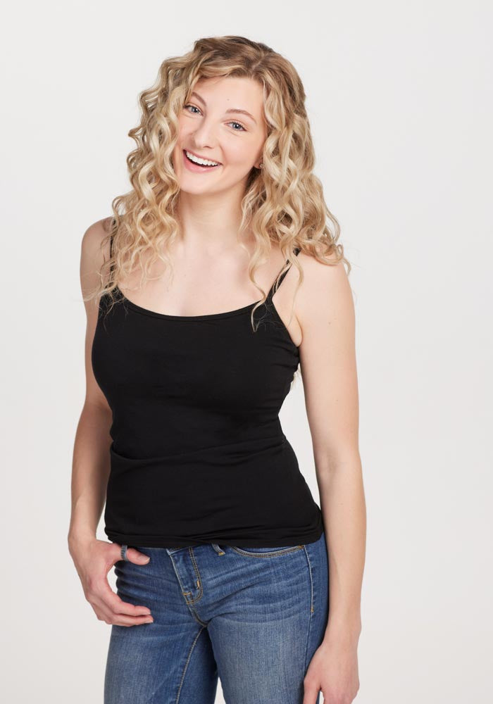 A smiling person with long, curly blonde hair is wearing the Teagen - Cami With Shelf Bra in black by Woolx and blue jeans. They are standing against a plain white background.