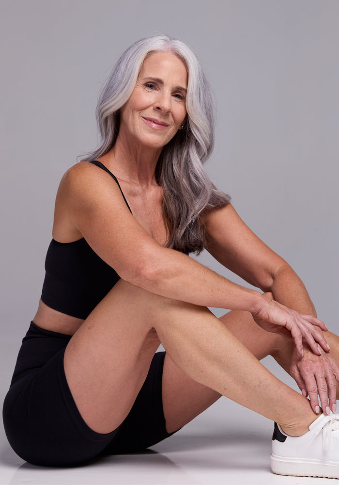 A woman with long gray hair sits on the floor, smiling at the camera. She is wearing a black sports bra and Woolx Marley Shapewear Shorts paired with white sneakers. Her left leg is bent, and her elbow rests on her knee. The background is a plain gray.