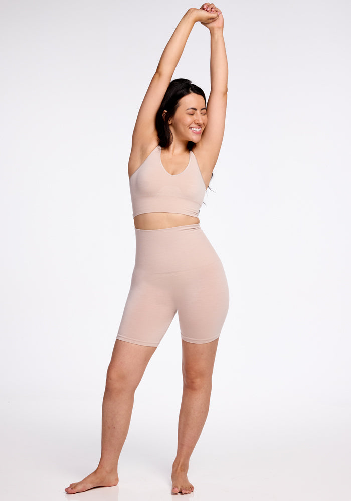 A woman stands on one leg, stretching with both arms raised above her head. She is smiling with her eyes closed and wearing a Woolx seamless construction matching beige sports bra and Marley Shapewear Shorts. The background is plain white.