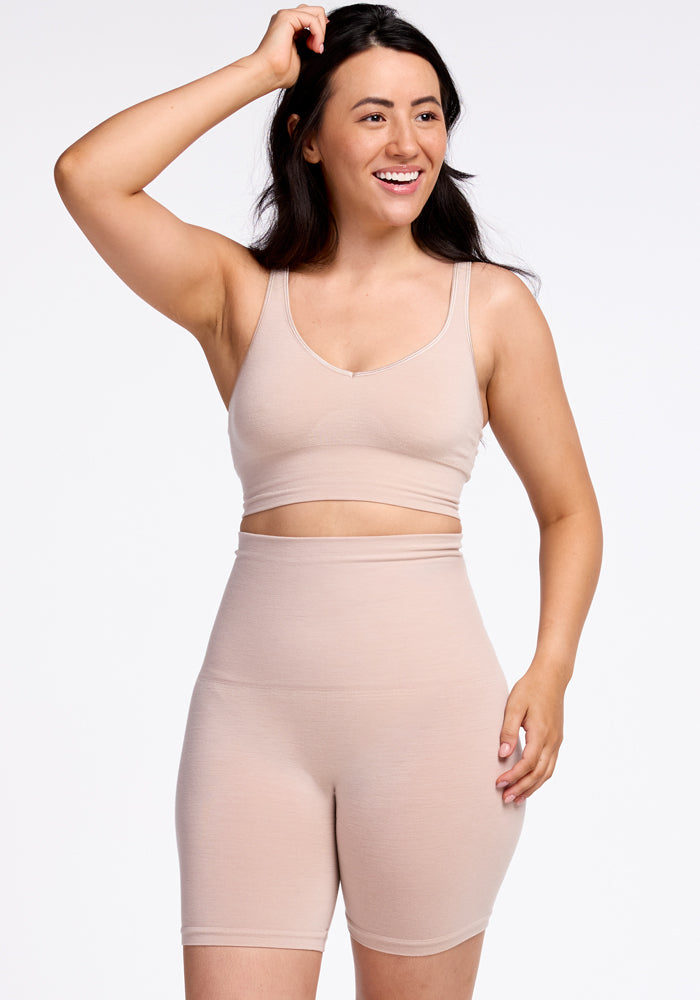 A person with long dark hair smiles and looks to the side while touching their head. They are wearing a matching beige workout outfit made of seamless construction, consisting of a crop top and Marley Shapewear Shorts from Woolx. The background is white, giving the image a clean and minimalistic look. 