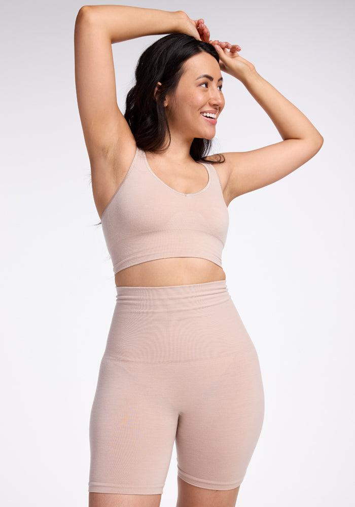 A person with long dark hair smiles while posing with their arms raised. They are wearing a matching beige Woolx sports bra and high-waisted Marley Shapewear Shorts made from merino wool, providing gentle compression against a plain white background.