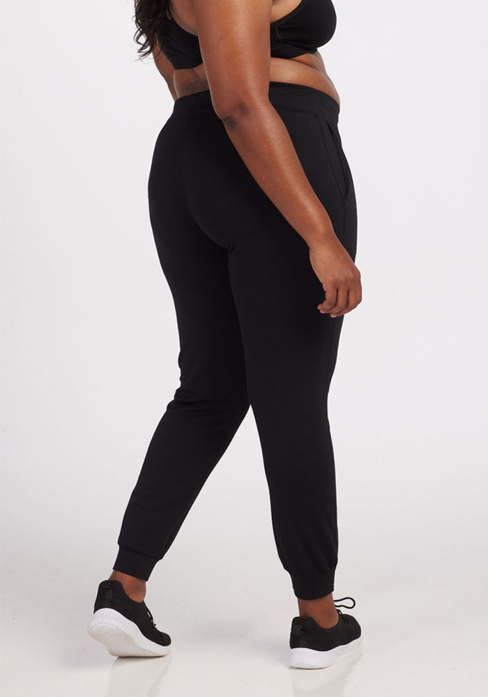 A person wearing the Woolx Lola Jogger in black, crafted from recycled polyester, stands with their back turned. They also sport sleek black sneakers with white soles against a plain white backdrop.