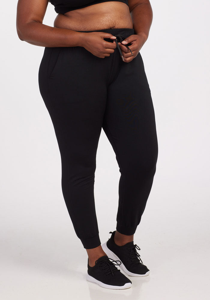 A person dressed in a black top and eco-friendly Lola Joggers by Woolx, crafted from recycled polyester, stands against a plain backdrop. Their hands are adjusting the waistband of the joggers. They complement their outfit with sleek black sneakers featuring white soles.