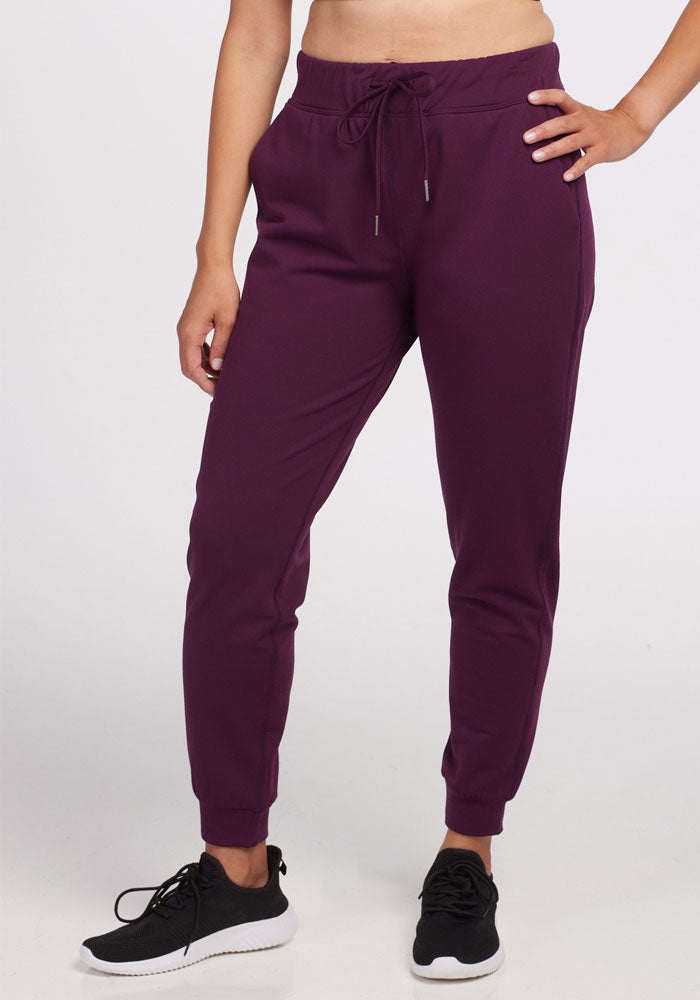 A person wearing burgundy Lola Jogger - Winter Bloom by Woolx and black sneakers stands against a plain white background. Made from recycled polyester, these joggers feature an adjustable drawstring waist, while the person's arms rest by their sides.