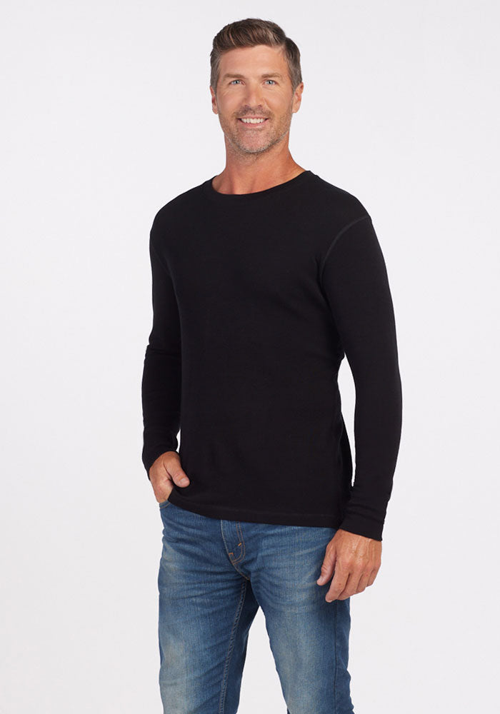 A man standing against a plain white background is smiling, wearing the Explorer Base Layer - Black from Woolx and blue jeans. His left hand is in his pocket, and his right hand is relaxed by his side.