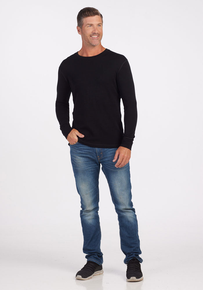 A person stands against a plain white background, dressed in a black Explorer Base Layer from Woolx, blue jeans, and black sneakers. Their right hand is in their pocket, and they are smiling while looking slightly to the left.