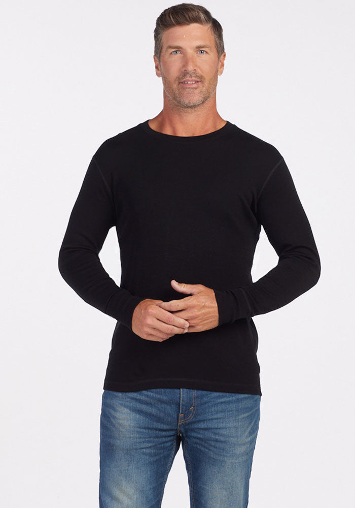 A man with short, slightly wavy hair is standing against a plain white background. He is wearing a black long-sleeve Explorer Base Layer from Woolx and blue jeans. He has a neutral expression and is facing the camera, with his hands loosely clasped in front of him.