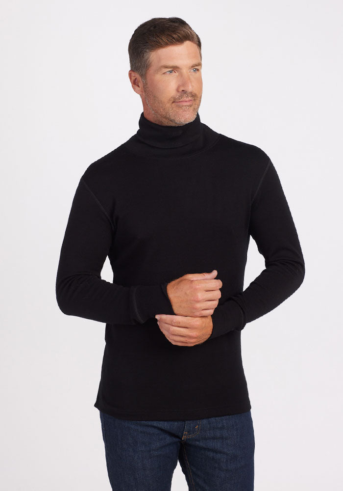 A person stands against a plain white background, wearing a Woolx Prescott Turtleneck in black made of luxurious Australian Merino wool and blue jeans. They have short brown hair and are casually holding one hand with the other in front of themselves while looking slightly to the side.