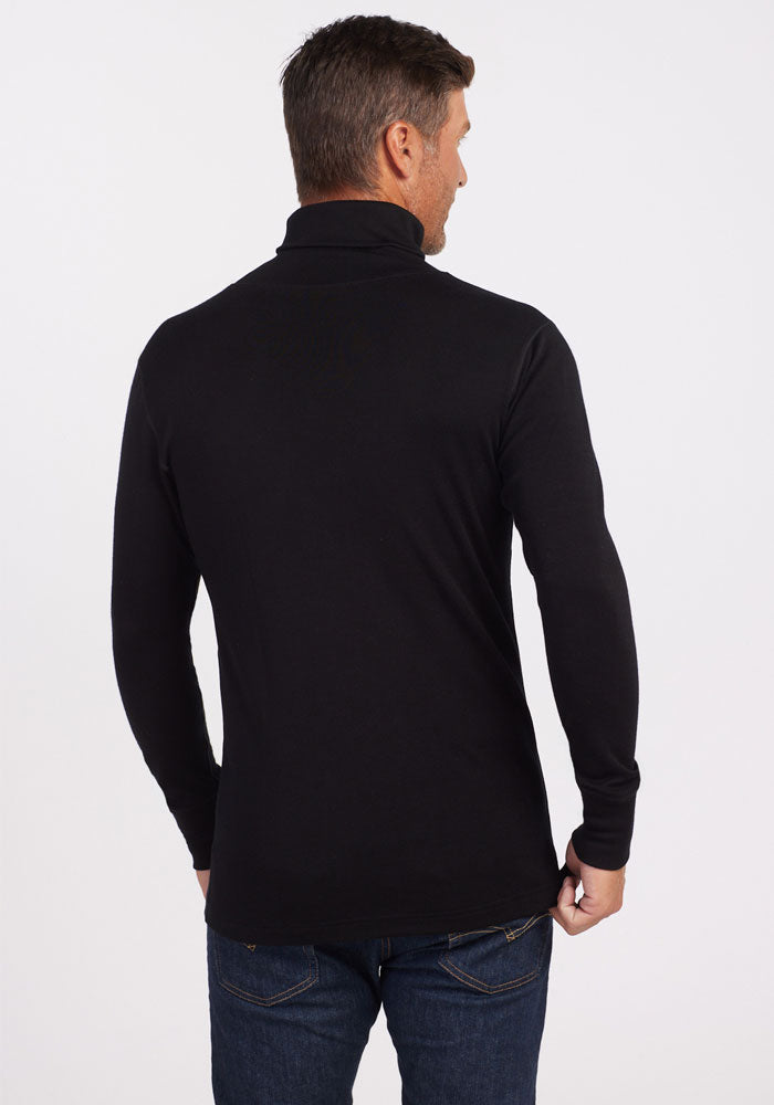 A man wearing a black Woolx Prescott Turtleneck, crafted from fine Australian Merino Wool, stands with his back to the camera against a plain white background. His hands rest near his hips as he slightly gazes over his left shoulder.