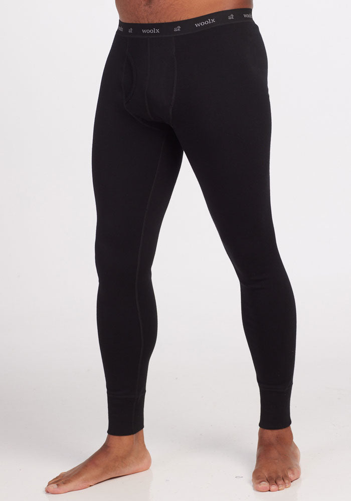 A person wearing Woolx's Backcountry Leggings in black, with "Woolx" branded on the waistband. These form-fitting Merino base layer bottoms extend down to the ankles and are ideal for cold weather adventures, featuring luxurious Australian Merino Wool. The person is standing barefoot on a plain white background. 