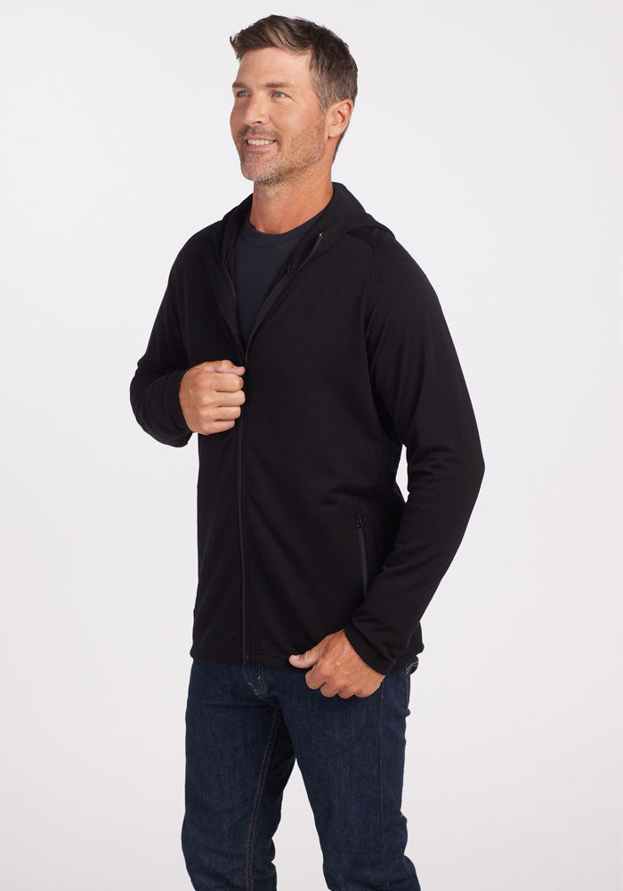 A man wearing a Woolx Boulder Hoodie - Black over a black shirt stands against a plain white background. He is slightly smiling and holding the zipper of his hoodie with one hand, while the other hand rests by his side. His casual style is completed with dark blue jeans.