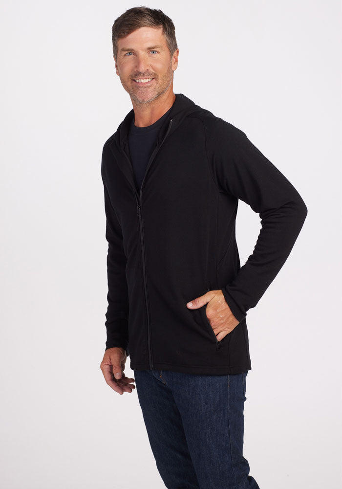 A smiling man with short brown hair and a beard is donning a Woolx Boulder Hoodie - Black over a dark shirt and blue jeans in a casual style. He stands against a plain white background with his left hand in his jacket pocket.