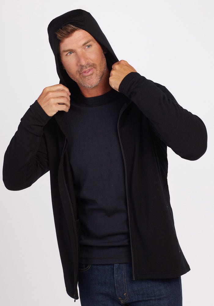 A person with short hair adjusts the hood of their Woolx Boulder Hoodie in black, crafted from Australian Merino Wool. They are also wearing a dark shirt underneath and dark blue jeans against a plain white background.