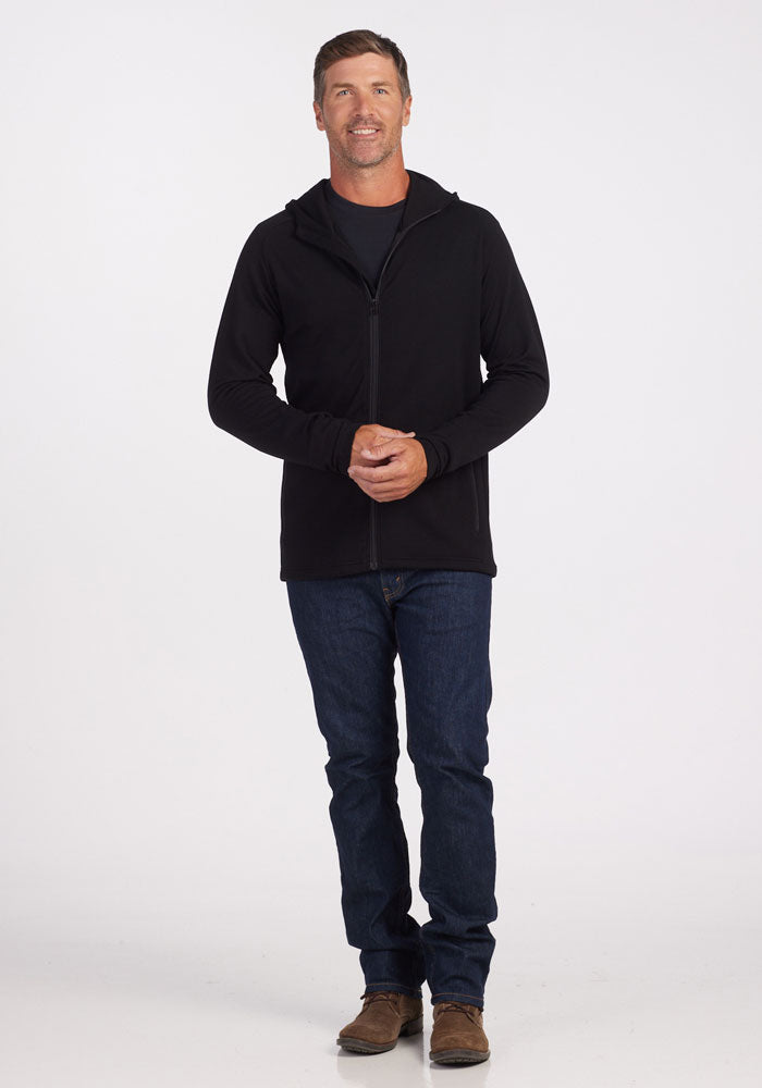 A man stands against a plain white background, wearing the Woolx Boulder Hoodie in black, crafted from Australian Merino Wool, over a dark T-shirt, blue jeans, and brown shoes. He has short brown hair and a slight smile, with his hands clasped in front of him.