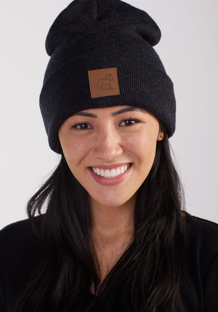 A person with long dark hair is smiling while wearing the Woolx Baylor Beanie in Carbon Black, which features a brown patch with a simple line drawing of an animal. They are dressed in a black top and posed against a plain light background.