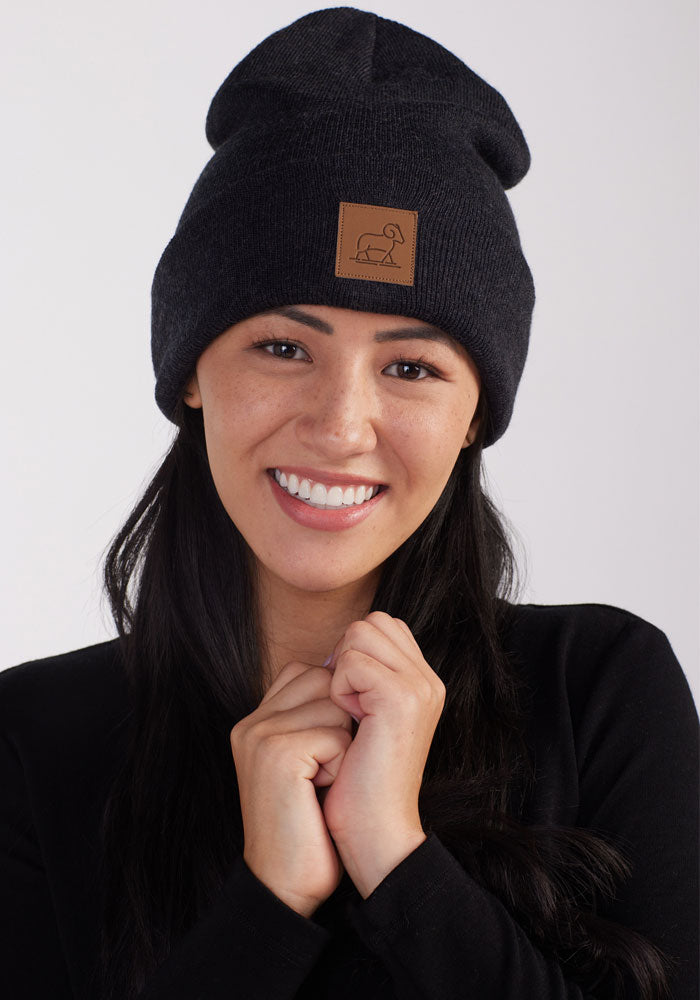 A woman with long, dark hair is smiling and looking directly at the camera. She is wearing a Woolx Baylor Beanie in Carbon Black, adorned with a brown patch featuring an animal logo. Dressed in a black top, she has her hands clasped near her face against a plain white background.