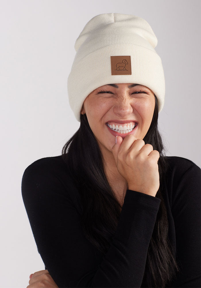 A person with long dark hair is smiling broadly with eyes closed, wearing a white Baylor Beanie - Sweet Cream by Woolx featuring a brown leather patch on the front and a black shirt. Their right hand is brought up to their mouth, and they have a joyful expression.