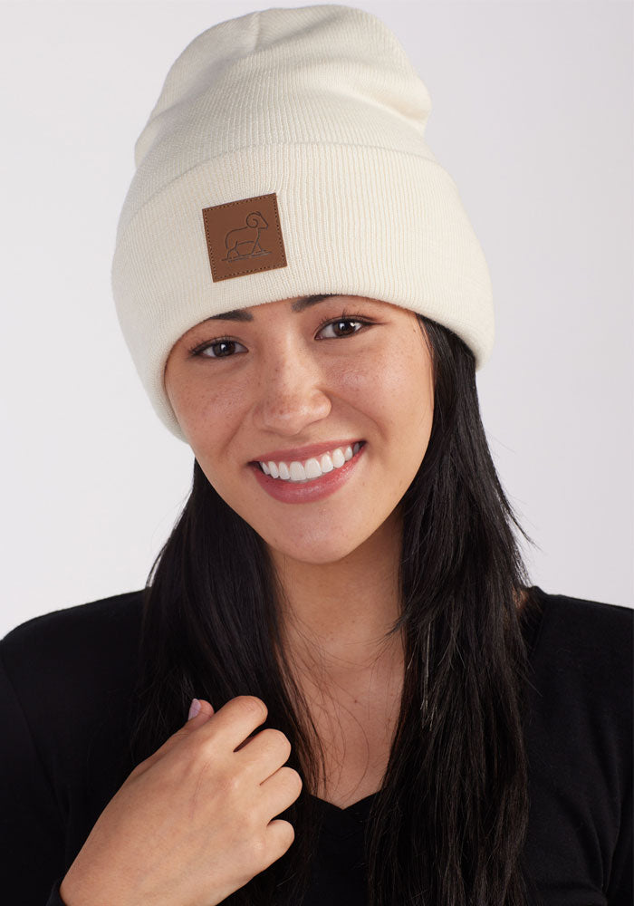 A smiling person with long black hair is wearing a white Baylor Beanie in Sweet Cream Merino wool by Woolx. The beanie features a brown leather patch with a simple line drawing on the front. They are dressed in a black top and are standing against a plain, light background.