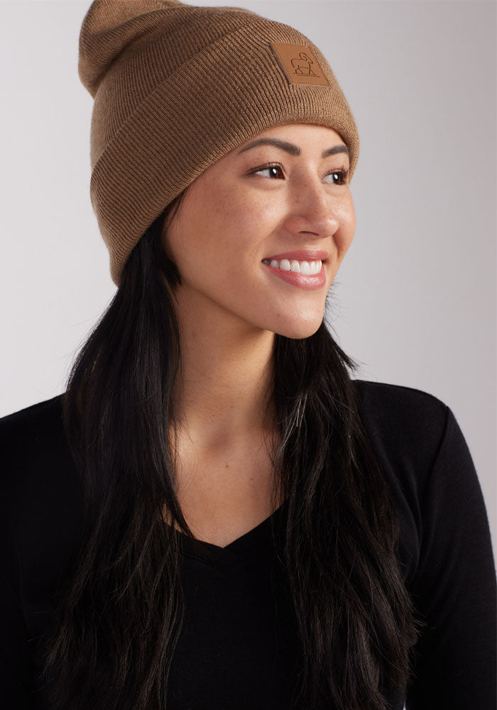 A person with long black hair is smiling while looking to the side. They are wearing a camel-colored Baylor Beanie from Woolx, featuring a small rectangular patch on the front, and a black V-neck top. The background is plain and light-colored.