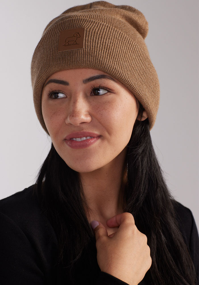 A person with long dark hair wearing a camel Baylor Beanie by Woolx and a black top looks off to the side with a slight smile. The beanie has a square patch on the front. The person has a hand lightly touching their chest. The background is plain and light-colored.