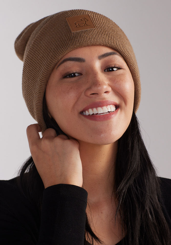 A person with long dark hair smiles while wearing a camel Baylor Beanie by Woolx. They have their hand touching the beanie near their face. The background is plain and light-colored.