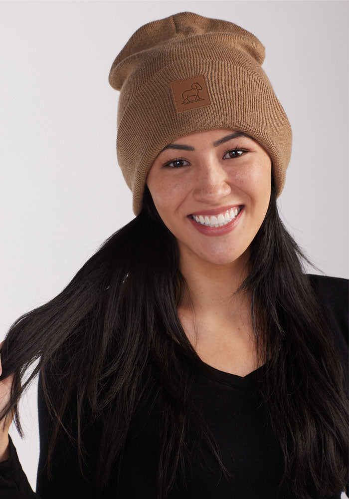 A person with long dark hair is smiling and holding strands of their hair. They are wearing a Camel Baylor Beanie by Woolx, which features a small leather patch, and a black top. The background is plain white.