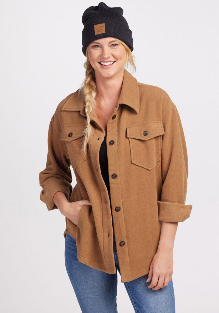 A woman with a braided hairstyle is smiling while wearing a Carbon Black Baylor Beanie by Woolx, a brown button-up shirt with rolled-up sleeves, and blue jeans. She has one hand in her pocket as she stands against a plain background.