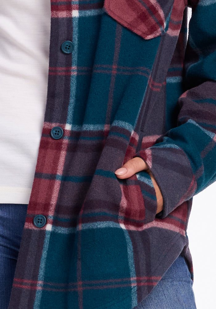 A close-up captures a person wearing the Sawyer Shacket by Woolx, featuring a teal, red, and grey plaid design. The shacket is partially buttoned over a white shirt, with the person's right hand tucked in their pocket and paired with blue jeans. The image highlights the distinct plaid pattern and texture of the Sawyer Shacket.