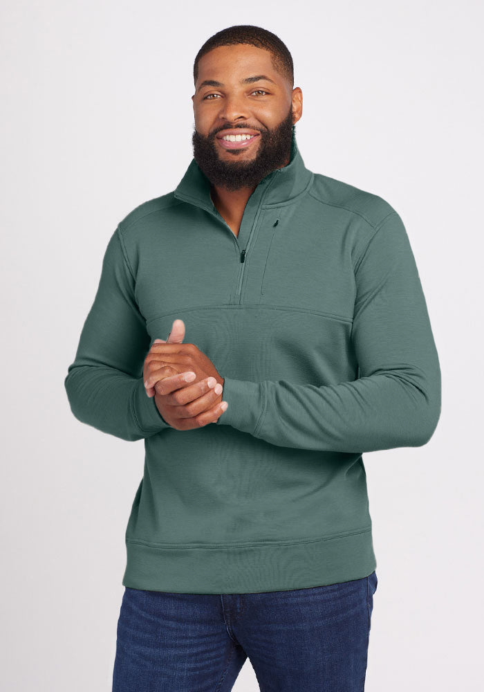 A bearded man smiles as he wears the Wool Aire™ Mason pullover in Duck Green from Woolx, paired with blue jeans. He stands against a plain white background with his hands clasped together in front of him. 