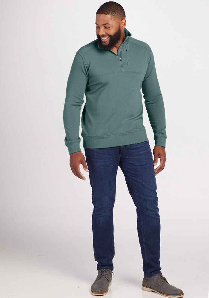 A man smiles while looking down, dressed in the Woolx Wool Aire™ Mason sweater in Duck Green. He pairs it with dark blue jeans and gray shoes, set against a plain white background.