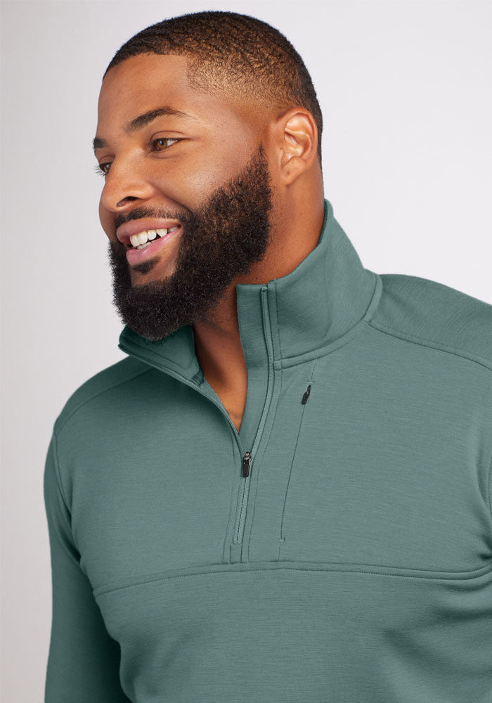 A man, smiling, is wearing the Woolx Wool Aire™ Mason - Duck Green pullover. This high-collared zip-up is crafted from Midweight WoolAire™ Fabric and features a convenient pocket. It looks comfortable, boasting a casual fit against the neutral gray background that emphasizes its details.