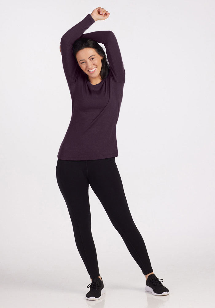 A person with long dark hair is standing, smiling and stretching their arms overhead with one leg slightly bent. They are wearing the versatile Kenzie Scoop Neck top from Woolx, black leggings, and black shoes with white soles. The background is white and plain.