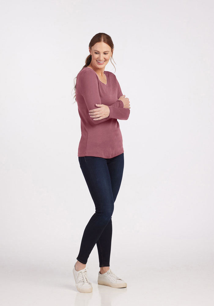 A woman with long red hair stands smiling with arms crossed, wearing the versatile Woolx Kenzie Scoop Neck top in mauve, dark blue skinny jeans, and white sneakers. She is posed against a plain white background, exemplifying a perfect blend of business casual elegance.