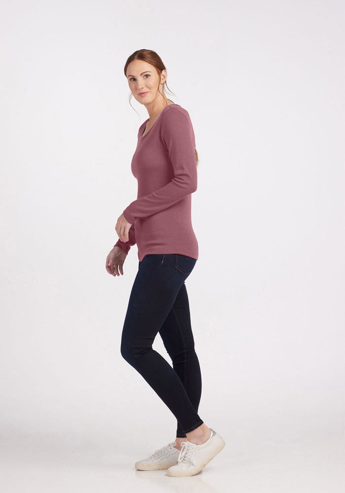 A woman with long, reddish hair is standing and looking at the camera. She is wearing the Kenzie Scoop Neck from Woolx in mauve, a long-sleeved, Merino wool top that adds a touch of business casual elegance. She has paired it with dark skinny jeans and white sneakers. The background is a plain, light grey color.