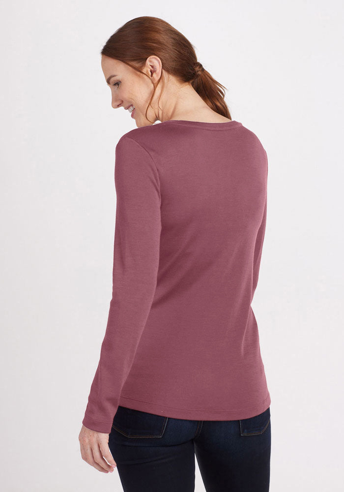 A woman with long, reddish-brown hair is standing against a plain background, turning slightly to her left. She is wearing a long-sleeved, dark pink Kenzie Scoop Neck top from Woolx and dark blue jeans. She is smiling and looking downwards, embodying a perfect business casual look.