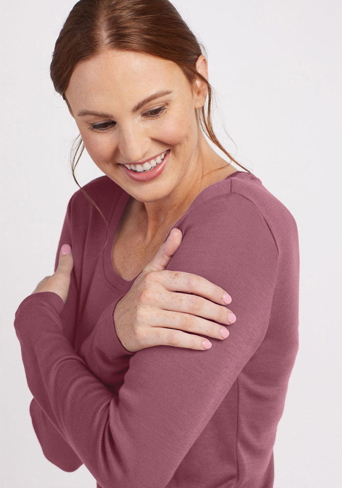 A smiling person with long red hair is wearing the versatile Woolx Kenzie Scoop Neck in pink. They are looking down and to their right, gently hugging their shoulders with both arms. The background is plain and light-colored, highlighting the elegance of the Australian Merino Wool shirt.