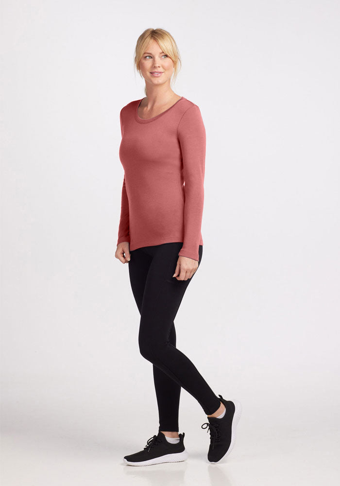 A woman is standing and posing against a plain background. She is wearing the Woolx Kenzie Scoop Neck, a long-sleeved, fitted pink top made of Merino wool, black leggings, and black athletic shoes with white soles. She has blonde hair styled in a ponytail and has a relaxed, cheerful expression.