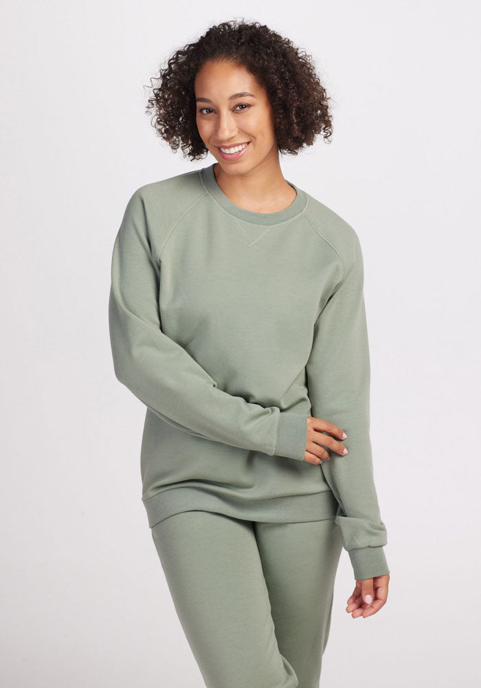 A person with curly hair smiles while wearing a sustainable Wool Aire™ Bailey sweatshirt and matching pants from Woolx. They stand against a plain white background, one hand on their hip and the other resting on their leg.