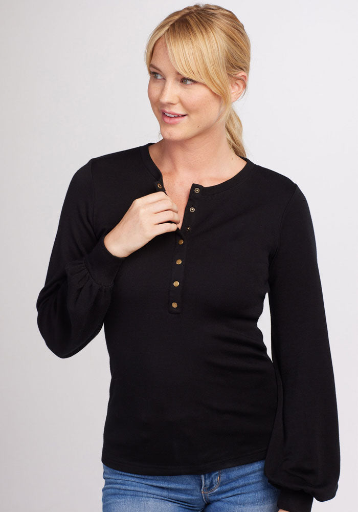 A person with blonde hair and a light complexion is wearing the Charlotte Puff Sleeve Top - Black from Woolx and light blue jeans. They are standing against a plain light background, looking to the side, and smiling slightly.