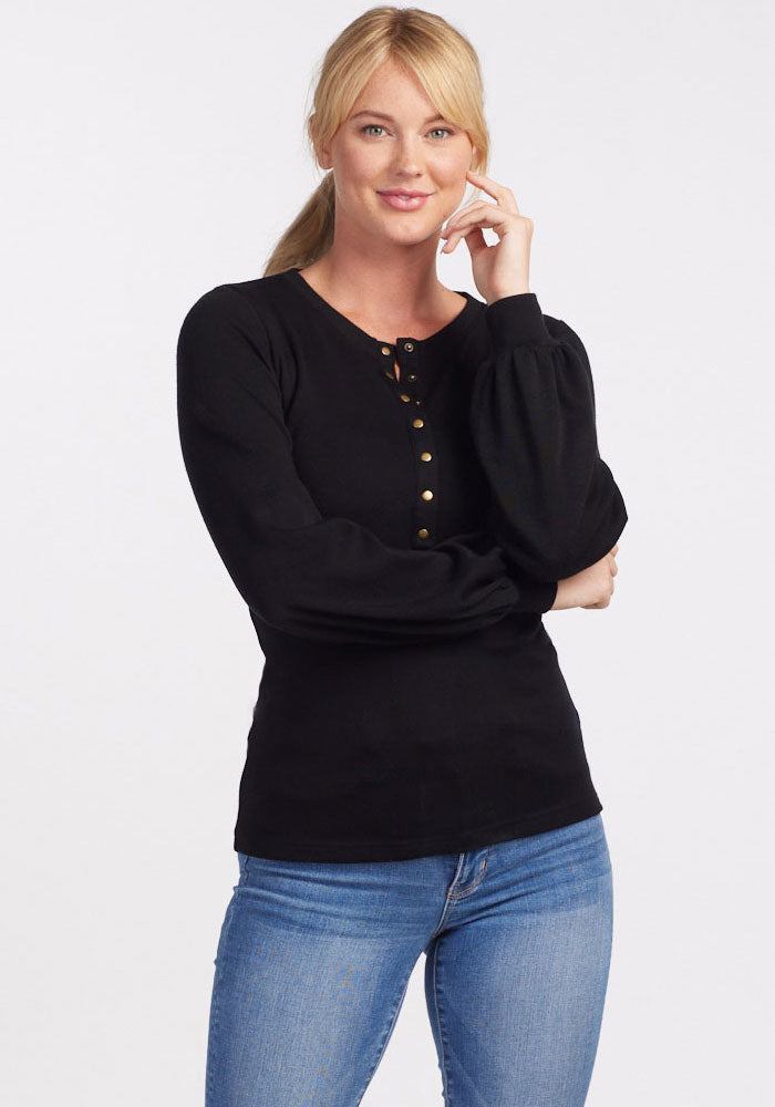 A woman with blonde hair is standing against a plain background, wearing the Charlotte Puff Sleeve Top - Black from Woolx and blue jeans. She is smiling, her right hand touching her face, and her left arm crossed in front of her.