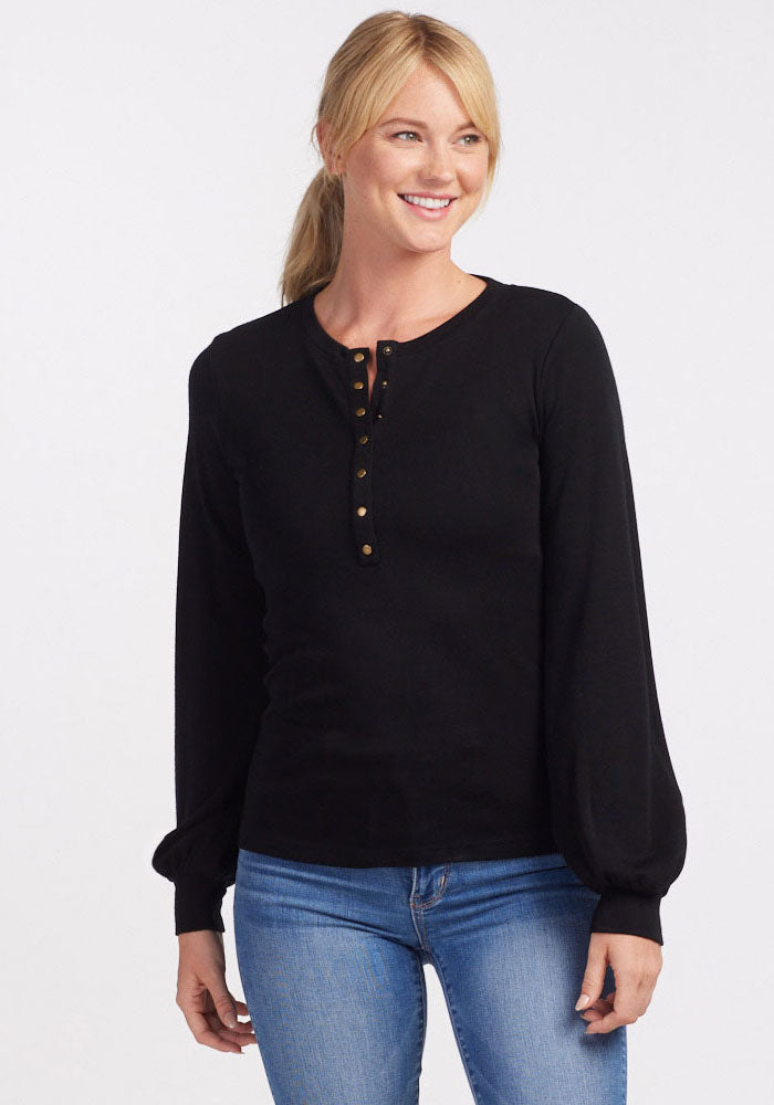 A smiling person with blond hair in a ponytail is wearing a long-sleeved Charlotte Puff Sleeve Top - Black by Woolx and blue jeans. The background is plain white. 