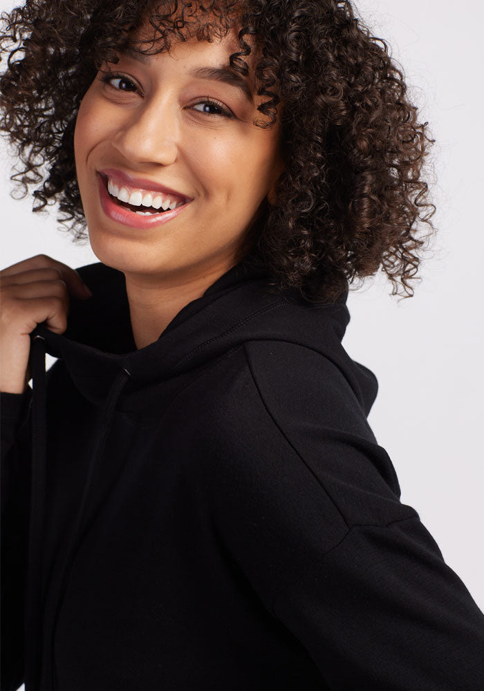 A person with curly hair is smiling warmly while holding the collar of a cozy Callie Hoodie - Black by Woolx. They are looking directly at the camera and appear very cheerful. The plain, light-colored background highlights their joyful expression and casual street style top.