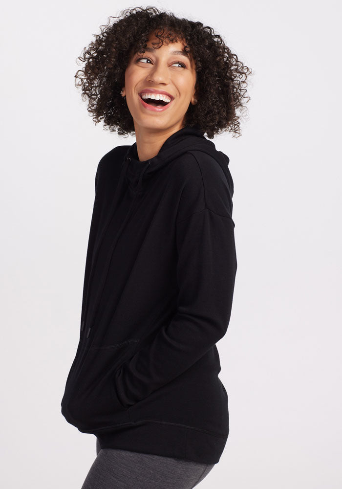 A person with curly hair is smiling and looking off to the side. They are wearing a cozy, warm Callie Hoodie in black from Woolx, with hands pocketed and gray leggings. The background is plain white.