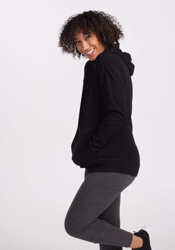 A person with curly hair is smiling and looking over their shoulder while wearing the Woolx Callie Hoodie in black, along with gray leggings and black sneakers. They have their hands in the hoodie pockets and are standing on one leg against a plain white background.