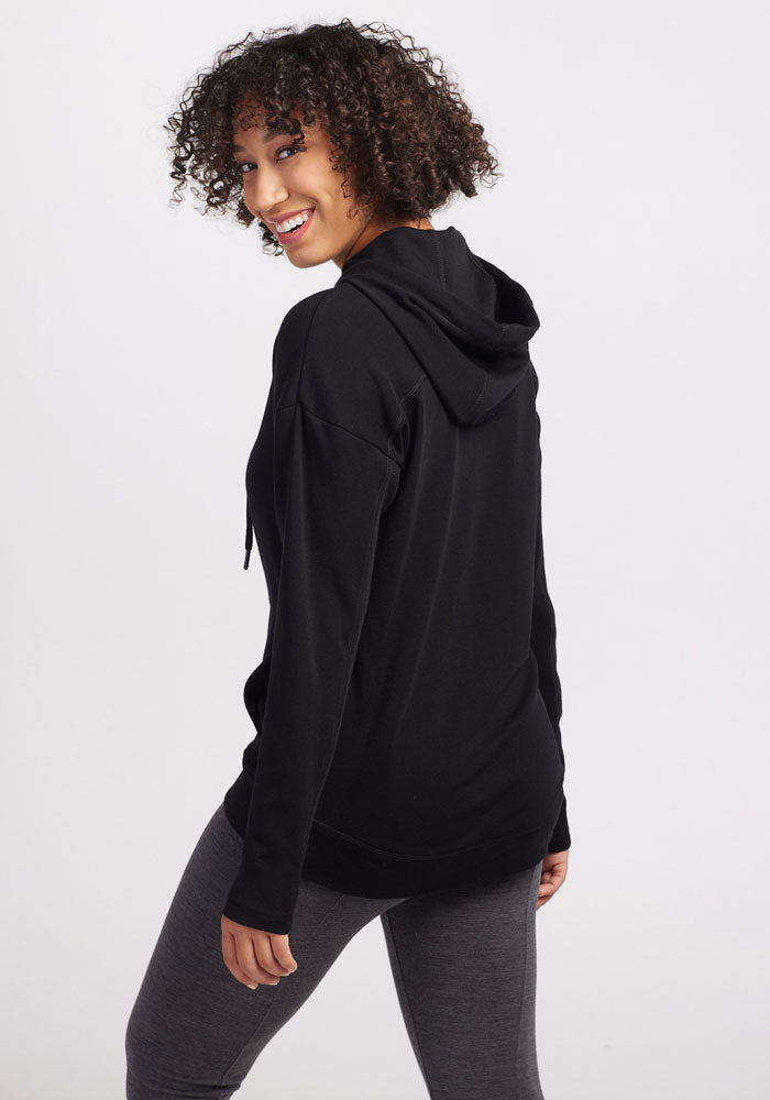 A person with curly hair is looking back and smiling while wearing a cozy Callie Hoodie - Black by Woolx and gray leggings, showcasing effortless street style against a plain white background.