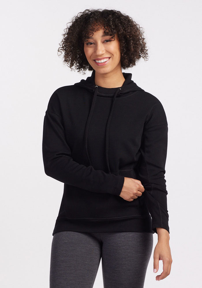 A person with curly hair is smiling while wearing a cozy Callie Hoodie - Black from Woolx and gray leggings. They are standing against a plain white background with one hand touching their hip and the other resting on their stomach. 