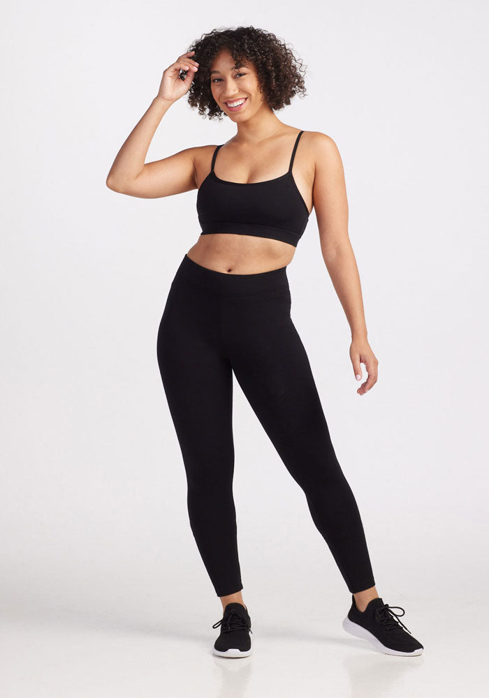 A woman with curly hair is smiling and posing in a black sports bra, Woolx Stella Leggings - Black, and black sneakers. She stands against a plain white background, with one hand touching her hair and the other resting by her side.